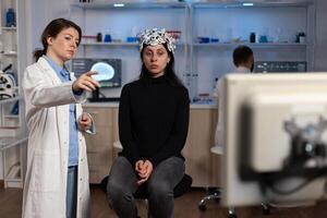 Neurologist doctor showing monitor with brain activity to woman patient with eeg scanner on head during neuroscience experiment in medical lab. Scientist physician analyzing nervous system evolution photo