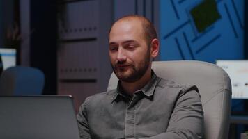 retrato do Exausta viciado em trabalho homem Gerente digitando financeiro estratégia usando computador portátil computador enquanto sentado às escrivaninha mesa dentro o negócio companhia escritório. cansado homem de negocios olhando para dentro Câmera atrasado às noite video