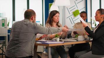 Young diverse team of workers talking, looking at documents and analyzing graphs data sitting at workplace desk in start up company. Professional colleagues team discussing marketing project. video