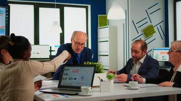 Senior businessman team leader brainstorming with colleague using paperwork showing ideas working in office. African woman using laptop in meeting broadroom, negotiations speaking with clients video