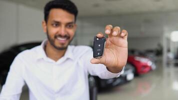 Positive indian male smiling for camera and demonstrating keys while standing near new vehicle in showroom video