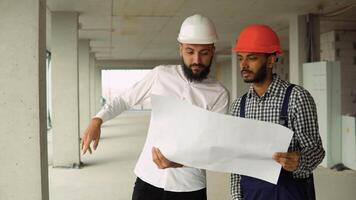 Arab architect is explaining to indian builder the plan of construction. Two young male men engineers wearing helmets holding a blueprint of the building and working interacting at construction site video