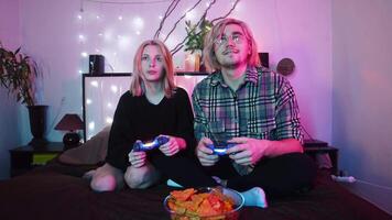 Young man and a woman are playing a game on a console in their room, illuminated by a bright neon light. They win the game and congratulate each other, giving each other a high five. video