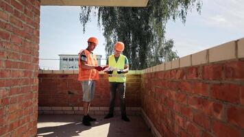 Two engineers wearing safety helmets are having a discussion on a construction site. Construction of a house. Engineers and architects review the construction plan. video