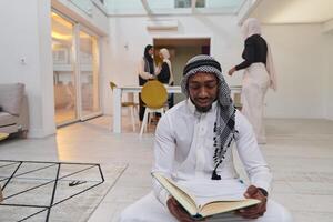 In the sacred month of Ramadan, an African American Muslim man engrossed in reading the Holy Quran is surrounded by a backdrop where three hijab-wearing women prepare and serve food for iftar photo