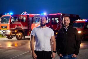 Group of firefighters, dressed in civilian clothing, stand in front of fire trucks during the night, showcasing a moment of camaraderie and unity among the team as they reflect on their duties and the photo