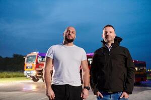 Group of firefighters, dressed in civilian clothing, stand in front of fire trucks during the night, showcasing a moment of camaraderie and unity among the team as they reflect on their duties and the photo
