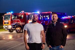 Group of firefighters, dressed in civilian clothing, stand in front of fire trucks during the night, showcasing a moment of camaraderie and unity among the team as they reflect on their duties and the photo