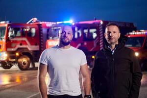 Group of firefighters, dressed in civilian clothing, stand in front of fire trucks during the night, showcasing a moment of camaraderie and unity among the team as they reflect on their duties and the photo