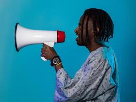 African American man dons traditional attire, passionately utilizing a megaphone against a striking blue background, symbolizing his vocal and cultural empowerment in the pursuit of social justice and photo