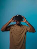 In a futuristic visual, an African American man stands isolated against a striking blue backdrop, adorned with VR glasses that transport him into a cutting-edge virtual reality experience, merging photo