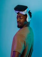 In a futuristic visual, an African American man stands isolated against a striking blue backdrop, adorned with VR glasses that transport him into a cutting-edge virtual reality experience, merging photo