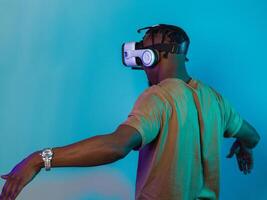 African American man dons VR glasses, engrossed in a virtual reality simulation, gesturing with hands, against a striking blue background, highlighting the fusion of cutting-edge technology and photo