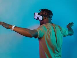 African American man dons VR glasses, engrossed in a virtual reality simulation, gesturing with hands, against a striking blue background, highlighting the fusion of cutting-edge technology and photo