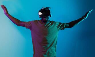 African American man dons VR glasses, engrossed in a virtual reality simulation, gesturing with hands, against a striking blue background, highlighting the fusion of cutting-edge technology and photo