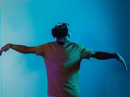 African American man dons VR glasses, engrossed in a virtual reality simulation, gesturing with hands, against a striking blue background, highlighting the fusion of cutting-edge technology and photo