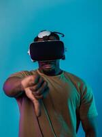 African American man, wearing virtual reality glasses, expresses dissatisfaction by pointing down with his hand while standing isolated against a blue background, portraying a modern critique within photo