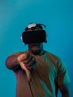 African American man, wearing virtual reality glasses, expresses dissatisfaction by pointing down with his hand while standing isolated against a blue background, portraying a modern critique within photo