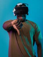 African American man, wearing virtual reality glasses, expresses dissatisfaction by pointing down with his hand while standing isolated against a blue background, portraying a modern critique within photo