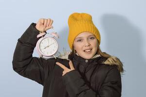Change to winter time for children. A girl in winter clothes points to an alarm clock with a dial photo