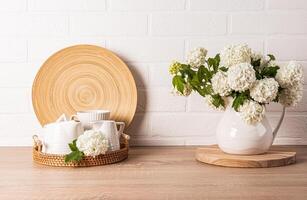 White ceramic tea set on round wicker tray on kitchen countertop. decorative flowers in jug. Kitchen background in light colors. Front view photo