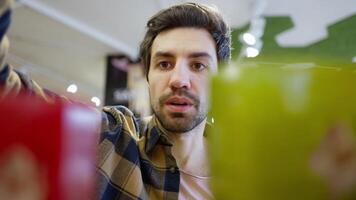 Close up a confident brunette guy with stubble in a checkered shirt chooses the products he needs while looking at a supermarket shelf video
