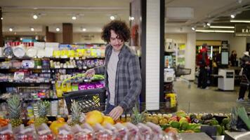 Happy guy with basket chooses citrus fruits in supermarket while shopping. Guy in a plaid shirt with curly hair in a grocery store video