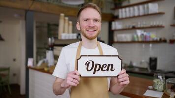 Portrait of a blond guy with a beard who holds in his hands a sign with an open sign in a cafe and poses video