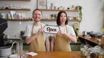 Portrait of a happy brunette girl in a yellow apron together with her colleague a blond guy raising a sign with an open sign while working in a cafe video