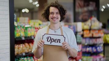 Portrait of a happy guy with black curly hair who smiles and holds a sign with the inscription open in his hands in a supermarket video