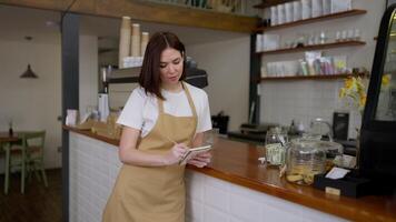 Portrait of a brunette waitress girl in a yellow apron who makes notes in her notebook near the cash register in a cafe video