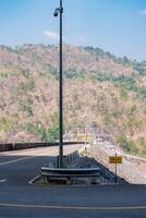 The scene of the road at the top of the dam, with the mountains in the background and a light pole. photo