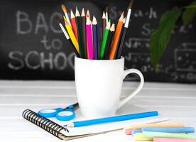 Colorful school stationery in a white mug on a wooden background. Blurred in the background is a blackboard with text. The concept of education. Selective focus. photo