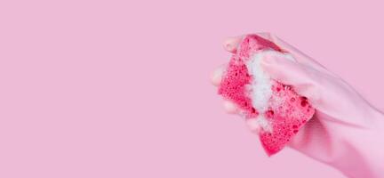 A women's hand in pink rubber glove holds a cleaning sponge with soapy foam on a pink background. Copy space. Banner. Selective focus. photo