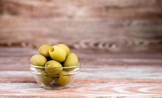 Green olives in a glass bowl on wooden background. Close-up. Copy space. photo