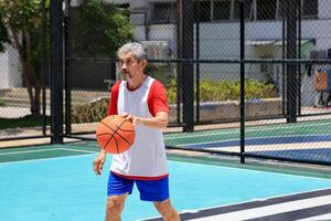 Senior Asian man in retirement age is playing basketball at outdoor court for healthy and fun activity to exercise and workout against aging illness photo