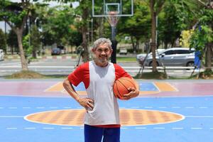 Portrait of senior Asian man in retirement age is playing basketball at outdoor court for healthy and fun activity to exercise and workout against aging illness photo