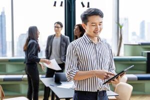 Portrait of young Asian businessman smiling with team having small meeting on the back while working in modern co-working space workplace for business and education concept photo
