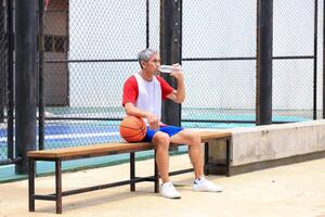Senior Asian man in retirement age is drinking water after playing basketball at outdoor court for healthy and fun activity to exercise and workout against aging illness photo