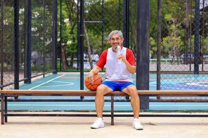 Senior Asian man in retirement age drinking water after playing basketball at outdoor court for healthy and fun activity to exercise and workout against aging illness photo