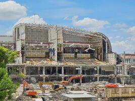 large building undergoing demolition. The structure is partially dismantled, revealing steel beams and fragmented concrete. Multiple heavy machines, including excavators, actively working on the site. photo
