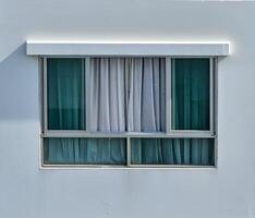 window set in a gray wall, featuring two glass panes with a metallic frame. Light gray curtains of a sheer fabric are partially drawn behind the glass, obscuring the view inside. Design photo