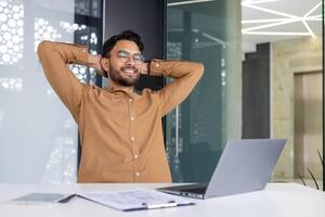 Smiling man relaxing at his desk with hands behind head, taking a break from work in a modern office environment photo