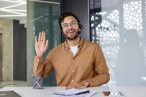 Happy man wearing a headset and waving while looking at the camera during a chat in a modern office setting. photo