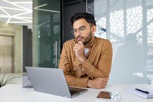 Young professional with glasses working on a laptop at a desk in a modern office. Considerate expression, focused on computer screen. photo