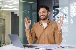 Young professional practicing yoga at his work desk in a modern office setting, showcasing relaxation and mental wellness. photo