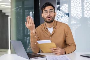 Confused man sitting at his desk holding an envelope and papers in a modern office setting, expressing uncertainty photo