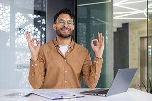 Young businessman practicing mindfulness meditation at his office desk with a laptop. Promoting workplace wellness and mental health. photo