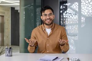 Smiling young Indian man sitting in the office at the desk and talking on a call, making an online recording, conducting training remotely. photo