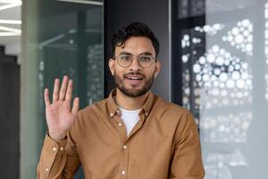Young man waving hand and looking at camera during a call in modern office. Professional communication, virtual meeting, and online conversation concept. photo